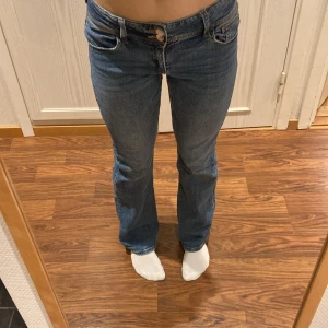 Blå bootcut jeans med låg midja - Snygga blå jeans med bootcut och låg midja. Bakfickorna har lock och knappdetaljer.