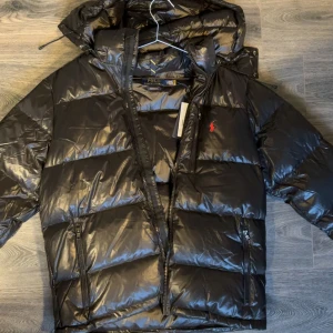 Svart pufferjacka från Polo Ralph Lauren - Svart pufferjacka från Polo Ralph Lauren med glansig finish och röd logobroderi på bröstet. Jackan har huva, dragkedja framtill och två sidofickor med dragkedja. Materialet är återvunnen polyester och jackan har en quiltad design som ger extra volym och värme.