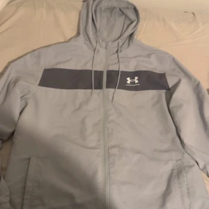 Grå vindjacka från Under Armour XL - Snygg grå vindjacka från Under Armour med mörkare grå panel över bröstet och vit logga. Jackan har huva med snörning, dragkedja framtill och praktiska sidofickor. Tillverkad i lätt polyester med meshfoder för extra komfort. Perfekt för aktiva dagar. Använda någon gång annars precis som ny nypris 800kr ungefär säljer för 650kr priset kan diskuteras fraktas inom 1-2 dagar 