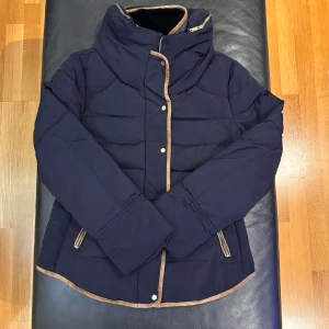 Mörkblå pufferjacka från Zara Basic - Snygg mörkblå pufferjacka från Zara Basic med beige kantdetaljer och hög krage. Jackan har dragkedja och tryckknappar framtill samt dragkedjor vid fickorna. Perfekt för kalla dagar och har en stilren, quiltad design.