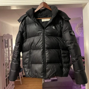 Svart pufferjacka Calvin Klein Jeans - Svart pufferjacka från Calvin Klein Jeans med stor huva och logga på bröstet och ärmen. Jackan har två sidofickor med dragkedja och är quiltad för extra värme. Perfekt för kalla dagar och har en clean, modern look.