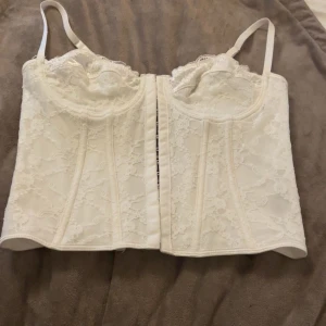 Vit spetskorsett med snörning - Säljer en vit korsett med vacker spets och justerbara axelband. Toppen har snörning i ryggen och formade kupor framtill för extra stöd. Perfekt för dig som vill ha en feminin och trendig look. Spetsdetaljerna ger en romantisk vibe och den ljusa färgen passar till många outfits. Pris kan diskuteras och köparen står för frakt 