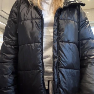 Svart pufferjacka med huva - Säljer en svart pufferjacka med huva och snörning. Jackan har quiltad design, dragkedja framtill och långa ärmar. Perfekt för dig som vill ha en varm och stilren jacka med loose passform. Materialet är syntet och jackan har en clean look.