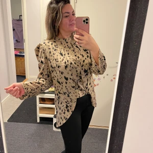 Beige blus med svart leopardmönster - Säljer en beige blus med svart leopardmönster och puffade axlar. Blusen har lång ärm, rund hals och är lite längre baktill. Materialet känns mjukt och har ett snyggt fall. Perfekt för dig som gillar att sticka ut med coola mönster.helt ny