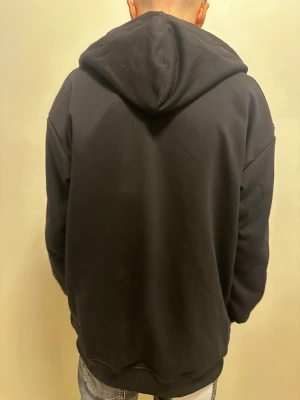 Svart Burberry hoodie med rutig huva - Svart hoodie från Burberry med dragkedja och stor broderad B-logga på bröstet. Insidan av huvan har klassiskt rutigt Burberry-mönster i svart, vitt och rött. Jackan har snörning i huvan och två stora fickor framtill. Perfekt för chill och streetwear.