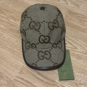 GG beige keps  - Snygg GG beige keps  med det klassiska GG-mönstret i brunt över hela kepsen. Kepsen har en böjd skärm med mörkbrun kant och justerbar rem i äkta skinn baktill. Perfekt för dig som vill ha en lyxig och trendig accessoar.