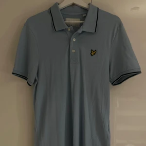 Ljusblå piké från Lyle & Scott - Fräsch ljusblå pikétröja från Lyle & Scott med klassisk krage och tre knappar framtill. Pikén har svart kant på kragen och ärmarna samt den ikoniska gula fågeln broderad på bröstet. Tillverkad i ekologisk bomull för extra skön känsla.