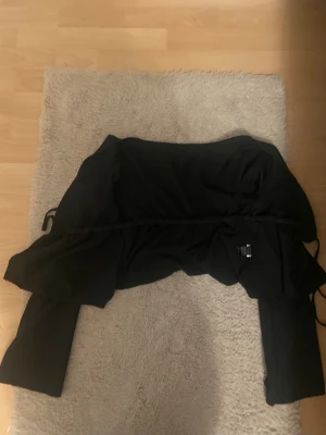 Svart omlott bolerotopp med knytband - Säljer en svart bolerotopp med långa ärmar och snygg omlott-design. Toppen har knytband i midjan för en justerbar passform och är croppad i modellen. Perfekt för att lyfta vilken outfit som helst med en stilren touch.