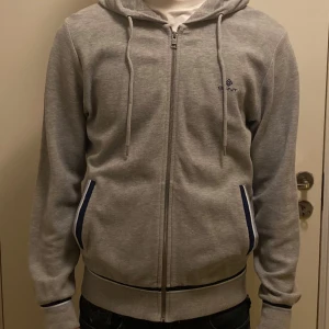 Grå hoodie från Gant med dragkedja - Snygg grå hoodie från Gant i storlek S. Tröjan har huva med snörning, hel dragkedja och två fickor med blå och vit kant. Ribbad nederkant och ärmslut med blå detalj. Perfekt för chill dagar och enkel att matcha med jeans.
