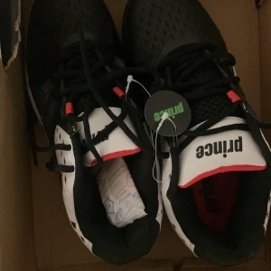 Svarta och vita Prince sneakers - Snygga sneakers från Prince i svart och vitt med röda detaljer. Ovandelen har mesh för bra andningsförmåga och sidorna är förstärkta med vita partier och svarta snören. Perfekta för dig som gillar sportig stil och vill sticka ut med en modern look.