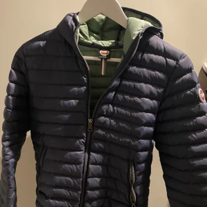 Mörkblå Colmar pufferjacka med huva - Säljer en mörkblå pufferjacka från Colmar med huva och grönt foder. Jackan har horisontella quiltade sömmar, två sidofickor med dragkedja och Colmar-logga på ärmen. Perfekt för dig som vill ha en snygg och funktionell jacka med cool streetkänsla.