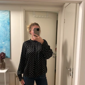 Svart prickig blus med puffärm - Säljer en svart blus med vita prickar och snygga puffärmar. Blusen har hög krage med rynkade detaljer och långa ärmar med breda muddar. Materialet är glansigt. 