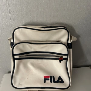 Vit axelväska från FILA - Retroinspirerad vit axelväska från FILA med svarta detaljer och logga framtill. Väskan har två dragkedjefack och justerbar axelrem. Perfekt för att bära med sig det viktigaste och ger en sportig vibe till outfiten.