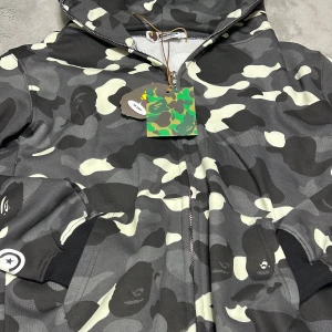 BAPE camo hoodie med shark-motiv - Säljer en BAPE hoodie med ikoniskt shark-motiv och broderade detaljer. Jackan har svart, grå och vit camouflage, dragkedja och huva. Stora bokstäver 'WGM' på sidan och feta grafiska prints. Perfekt för dig som vill sticka ut med streetwear.