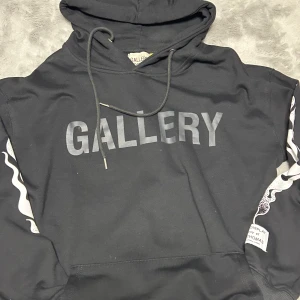 Svart hoodie från Gallery Dept. - Svart hoodie från Gallery Dept. med stor 'GALLERY' print på bröstet och coola vita grafiska detaljer längs ärmarna. Tröjan har huva med snörning och en klassisk känguruficka. Materialet är mjuk bomull och polyester, perfekt för en chill och stilren look.