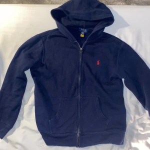 Mörkblå hoodie från Polo Ralph Lauren - Snygg mörkblå hoodie från Polo Ralph Lauren med klassisk röd logobrodyr på bröstet. Tröjan har huva, dragkedja framtill och två stora fickor. Mjuk insida i fleece och långärmad modell, perfekt för kyliga dagar. Storlek 14-16 år (xs) mycket bra skick inga synliga defekter. Hör gärna av er vid frågor!!