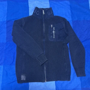 Marinblå stickad kofta Sail Racing - Säljer en marinblå stickad kofta från Sail Racing med ribbad struktur och hög krage med logotyp. Koftan har hel dragkedja framtill och en snygg bröstficka med dragkedja och Sail Racing-tryck. Perfekt för dig som gillar sportig och clean stil.