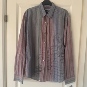 Randig skjorta från Desigual L - Unik skjorta från Desigual i storlek L med färgglada ränder i blått, rött, grönt och orange. Skjortan har lång ärm, klassisk krage och knappar framtill. Materialet är bomull och mönstret är randigt med coola handskrivna detaljer på framsidan.