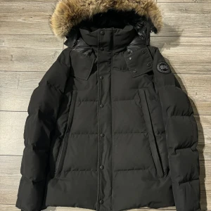 Svart dunjacka från Canada Goose - Säljer en svart dunjacka från Canada Goose med fet pälskant på huvan och ribbade muddar vid ärmslut. Jackan har lång ärm, flera fickor med knappstängning och klassisk patch på axeln. Perfekt för kalla vinterdagar och riktigt snygg siluett.