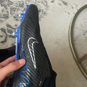 Nike Zoom fotbollsskor svart/blå - Säljer ett par svarta Nike Zoom fotbollsskor med blå detaljer och vit swoosh. Skorna har en strömlinjeformad design med platt sula och syntetmaterial för bästa grepp på planen. Perfekt för dig som vill ha snygga och funktionella skor till matchen.