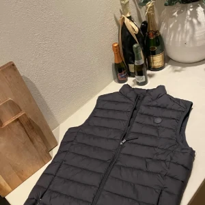 Svart dunväst med dragkedja - Snygg svart dunväst från Peak Performance med quiltad design och hög krage. Västen har en diskret logga på bröstet och praktisk dragkedja framtill. Perfekt att slänga över en hoodie eller tröja för extra värme under kyliga dagar.