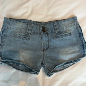 Blå jeansshorts med rå kant - Säljer ett par ljusblå jeansshorts med fransig rå kant och dubbla knappar framtill. Shortsen har klassiska fickor både fram (fejk) och bak samt hällor för bälte. Perfekta för varma dagar och har en snygg, avslappnad vibe. Storlek 31 