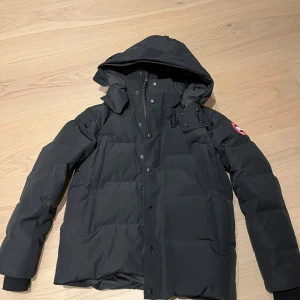 Svart dunjacka från Canada Goose - Svart dunjacka från Canada Goose med huva och klassisk logga på ärmen. Jackan har quiltad design, dragkedja och knappar framtill samt två stora fickor. Perfekt för kalla dagar och har en clean, modern look som funkar till det mesta. Använd 3 gånger.