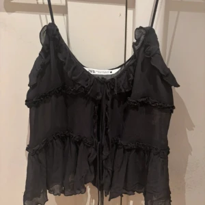 Svart volangig linnetopp från Zara - Supersöt svart linnetopp från Zara med tunna axelband och flera lager volanger. Toppen har en luftig och flowy känsla, perfekt för att skapa en cool och avslappnad look. Knytning framtill ger extra detalj och toppen är lätt transparent.