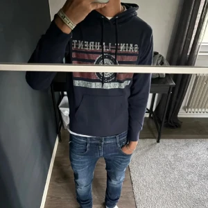 Mörkblå Saint Laurent Hoodie - Snygg mörkblå hoodie från Saint Laurent med tryck i vitt och rött framtill. Tröjan har huva med snörning, känguruficka och lång ärm. Materialet är mjuk bomull och passformen är normal. Perfekt för dig som gillar streetwear med exklusiv vibe. Kvitto, äkthetsbevis finns.