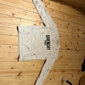 Vit Givenchy sweatshirt med hål - Säljer en vit sweatshirt från Givenchy med svart tryck 'GIVENCHY PARIS' på bröstet. Tröjan har lång ärm och är designad med slitna hål över hela plagget för en edgy look. Mjuk bomullskvalitet och klassisk rund hals.
