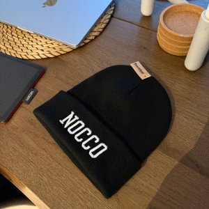 Svart NOCCO mössa med broderad logga - Svart mössa från NOCCO med vit broderad logotyp framtill. Klassisk ribbstickad design och uppvikt kant. Perfekt för kalla dagar och snygg att matcha med streetwear. Materialet är stickad syntet, vilket gör den både mjuk och varm.