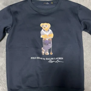 Mörkblå sweatshirt Polo Ralph Lauren - Snygg mörkblå sweatshirt från Polo Ralph Lauren med ikoniska Polo Bear-trycket på bröstet. Tröjan har rund hals och ribbade muddar. Perfekt för dig som gillar klassisk streetstyle med en twist. Materialet är mjuk bomull som känns skönt mot huden.