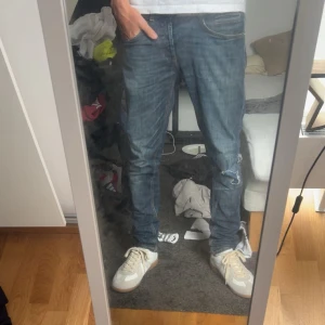 Blå jeans med slitningar - Säljer ett par blå jeans med snygga slitningar på ena benet. Jeansen har klassisk femficksdesign, normal passform och är tillverkade i jeansmaterial. Perfekta för en avslappnad och trendig look.
