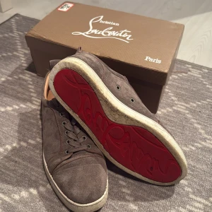 Grå vita loubs utan spikar - Snygga grå sneakers från Christian Louboutin i mocka med klassisk röd sula och rund tå. Skorna har snörning och en platt vit sula med signaturdetaljer undertill. Insidan är beige och de har en stilren, urban look som passar till jeans och hoodie.