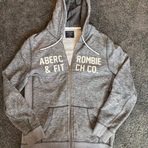 Grå hoodie Abercrombie & Fitch - Snygg grå hoodie från Abercrombie & Fitch i storlek S. Tröjan har vit broderad logga framtill, huva med vita snören och praktisk dragkedja. Tillverkad i mjuk bomullsmix med ribbade muddar och fickor framtill. Perfekt för chill dagar.
