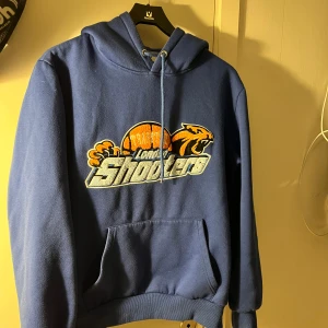 Blå hoodie Trapstar London Shooters - Blå hoodie från Trapstar med stort London Shooters-tryck i orange, grått och vitt på bröstet. Tröjan har huva med snörning och en klassisk magficka. Perfekt för dig som gillar streetwear och sportiga vibes.