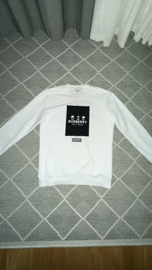 Vit sweatshirt från Burberry, M - Vit sweatshirt från Burberry med svart tryck framtill och klassisk rund hals. Tröjan är i mjuk bomullsmix med lite stretch för extra komfort. Långärmad och stilren design som lyfter din outfit med en lyxig touch.