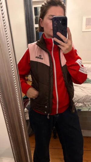 Adidas brun och rosa dunväst - Säljer en dunväst från Adidas i brunt med ljusrosa axelparti och svart Adidas-logga på bröstet. Västen har quiltad design, knappar framtill och en sportig look som passar perfekt till streetwear. Perfekt att slänga över en hoodie eller tröja.