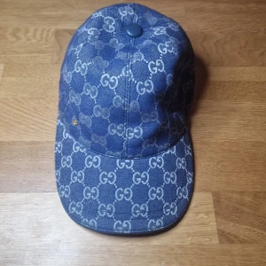 Blå Gucci keps med GG-mönster med låda - Snygg blå keps från Gucci med ikoniskt GG-mönster över hela kepsen. Justerbar rem baktill i blått läder med guldfärgad detalj. Materialet är bomull och kepsen har en klassisk böjd skärm. Perfekt för dig som vill ha en lyxig streetstyle-look.