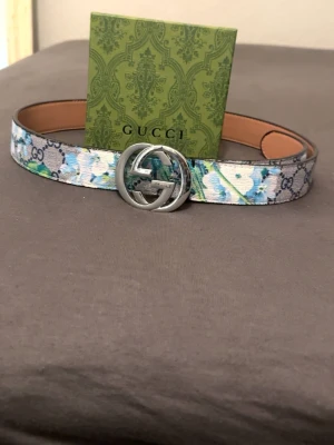 Gucci blommigt bälte med silverdetaljer - Säljer ett Gucci bälte med blommigt mönster i blått, grönt och vitt. Bältet har en stor, rund silverspänne med GG-logga och är gjort i skinn med canvas-yttersida. Perfekt statement piece för att lyfta din outfit.pris kan diskuteras
