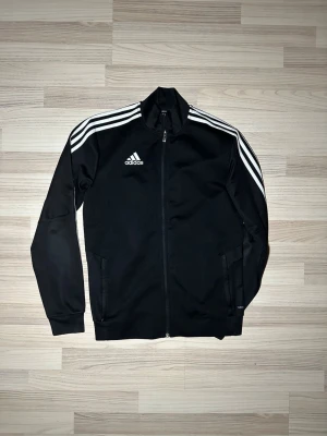 Svart Adidas vindjacka med vita ränder - Säljer en svart Adidas vindjacka med klassiska vita ränder längs ärmarna och Adidas-logga på bröstet. Jackan har hel drag soppa, ståkrage och två fickor framtill. Tillverkad i polyester och perfekt för sportiga outfits.
