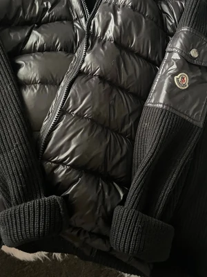Moncler Cardigan Svart - Säljer en svart dunväst från Moncler med quiltad front och stickade detaljer på sidor och krage. Västen har dragkedja framtill och en Moncler-logga på ärmen. Materialmix av polyester och stickat ger en schysst kontrast och modern look.