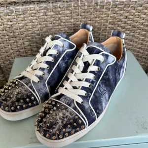 Christian Louboutin sneakers med nitar - Snygga sneakers från Christian Louboutin med mörkblått och grått mönster, vita snören och vita detaljer. Tåpartiet är täckt av silvriga nitar för en edgy look. Skorna har rund tå och platt sula, samt lyxig läderinsida.