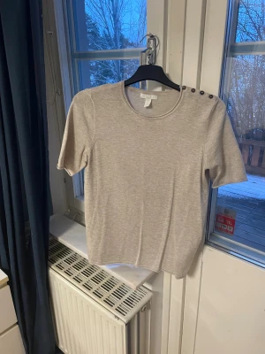 Beige stickad tröja med knappar H&M - Beige stickad tröja från H&M Basics i storlek S. Rund halsringning och korta ärmar. Snygga mörka knappar på ena axeln ger en extra detalj. Perfekt för dig som gillar enkel och stilren design med en twist.