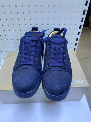 Mörkblå mockasneakers från Louboutin - Snygga mörkblå sneakers i mocka från Louboutin med klassisk röd sula och rund tå. Skorna har matchande mörkblå snörning och diskreta sömmar för en clean look. Perfekta för dig som gillar exklusiva detaljer och streetstyle.