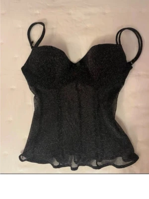Svart glittrig Intimissimi topp - Supersnygg svart korsettopp från Intimissimi med sopiga glitterdetaljer. Tunn mesh med lätt utsvängd nederkant och dubbla smala axelband. Perfekt för dig som vill sticka ut lite extra. Prislapp kvar, oanvänd. 