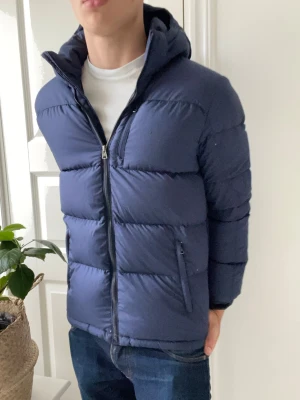 Ralph Lauren dun jacka  - Ralph Lauren dun jacka, en riktigt skön mörkblå Ralph Lauren dun jakca med skön luva som går att knäppa upp / av. Inga direkta defekter förutom att loggan har tappat lite av sin färg. Denna modellen av jacka är en barn version därför är loggan dit målad istället för den kändaste modellen fast sydd. Modellen som bär den är 180 cm 75 kg och den är på gränsen lite för liten. 🙏Xl i barn är ungefär en S i herr 
