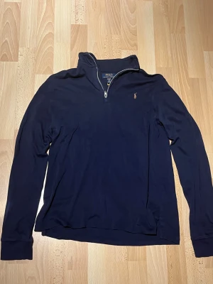 Marinblå half zip tröja Polo Ralph Lauren - Snygg marinblå långärmad tröja från Polo Ralph Lauren. Tröjan har en klassisk half zip-dragkedja och det ikoniska broderade Polo-logot på bröstet. Tillverkad i mjuk bomull, perfekt för höst- och vårväder.