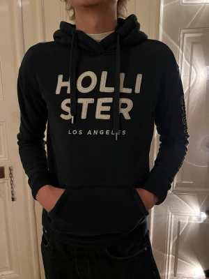 Hollister Hoodie - Storlek S - Fint Skick - Säljer Klassisk marinblå hoodie från Hollister med stor vit broderad logotyp "HOLLISTER" och "LOS ANGELES" under. Signatur "CALIFORNIA" text längs vänster ärm. En perfekt vardagsfavorit som passar till allt! Den är väldigt trendig nu så var snabb o handla!  Detaljer: Märke: Hollister California Storlek: S (Small) Färg: Svart med vit logotyp Modell: Pullover hoodie med dragsko Skick: Mycket gott - minimala tecken på användning