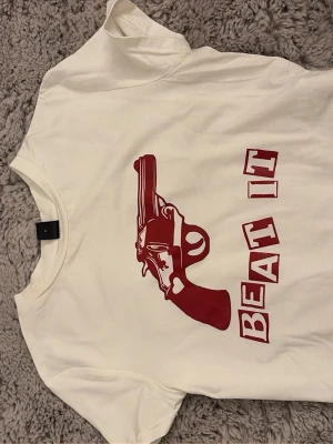 Vit t-shirt med röd pistolprint från SHEIN - Vit t-shirt från SHEIN i storlek S med ett stort rött tryck av en pistol och texten 'BEAT IT' på framsidan. Klassisk rund hals och korta ärmar. Tillverkad i mjuk bomull som känns skön mot huden.